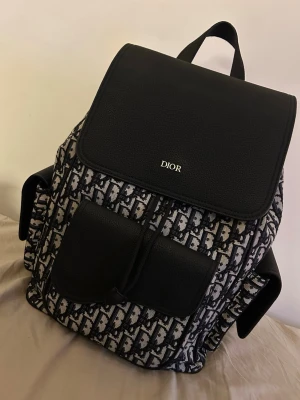 Dior ryggsäck i svart och monogram - Snygg ryggsäck från Dior med klassiskt svart och vitt monogrammönster. Väskan har lock och fickor i svart läder, samt justerbara axelremmar. Perfekt för dig som vill ha en lyxig och trendig accessoar med plats för allt du behöver.