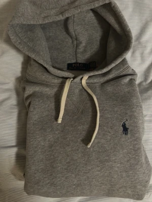 Grå hoodie från Polo Ralph Lauren - NY RALPH LAUREN HOODIE. Nypris 1699kr Mitt pris 899kr.  Broderad marinblå logga på bröstet. Mjuk bomullsblandning som är skön att ha på sig. Perfekt för en avslappnad och stilren look.