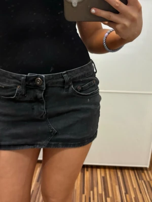 Svart jeanskjol med fickor - Snygg svart kort jeanskjol med klassiska fickor fram och bak samt bälteshällor. Kjolen har knapp- och dragkedjestängning framtill och en tight passform som sitter snyggt på kroppen. Perfekt att matcha med en enkel topp eller hoodie.