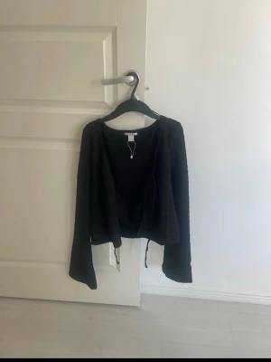 Svart omlottkofta med vida ärmar - Snygg svart omlottkofta med långa, vida ärmar och knytband i midjan. Perfekt att slänga över en topp för en avslappnad men trendig look. Enkel design som passar till mycket och ger en cool vibe och de finns som ett linne under så du behöver inte ha något under tröjan