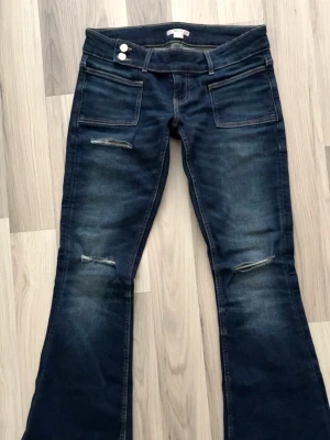 Mörkblå bootcut jeans från Gina 14+ - Säljer ett par mörkblå bootcut jeans med slitningar på låren och knäna. Jeansen har stora framfickor, dubbla knappar i midjan och en tvättad look. Perfekta för en avslappnad och trendig stil. Lite slitna längst ner då dem är lite nertrampade men annars väldigt fint skick💗🩷