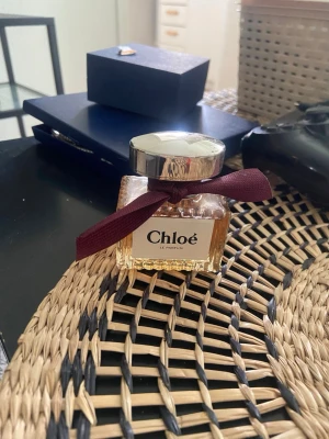 Chloé Le Parfum med vinrött band - Chloé Le Parfum i en elegant glasflaska med silverfärgat lock och ett vinrött tygband runt halsen. Flaskan har en klassisk, lyxig design och parfymen har en ljus bärnstensfärg. Perfekt för dig som gillar stilrena och exklusiva dofter.