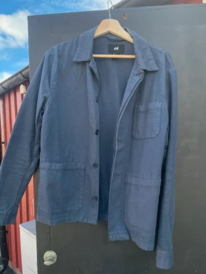 Blå overshirt från H&M - Stilren blå overshirt från H&M med klassisk krage och tre framfickor. Lätt och smidig modell i bomull, perfekt att slänga över en t-shirt. Enkel knäppning framtill och raka ärmar ger en avslappnad look. 48 men passar som small.