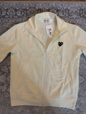 Gul zip-hoodie från Comme des Garçons - Snygg ljusgul hoodie med dragkedja från Comme des Garçons PLAY. Har det ikoniska svarta hjärtat med ögon på bröstet och en stor version på ryggen. Mjukt material och två fickor framtill. Perfekt för dig som gillar streetwear och vill sticka ut.