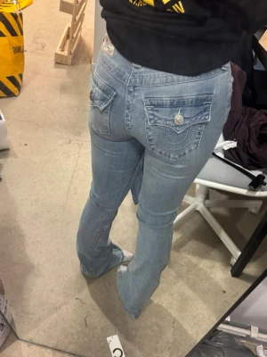 Ljusblå bootcut jeans från True Religion - Säljer ett par ljusblå bootcut jeans från True Religion med ikoniska bakfickor och silverfärgade knappar. Jeansen har låg midja och klassisk femficksdesign. Perfekta för dig som gillar en avslappnad men ändå trendig look.