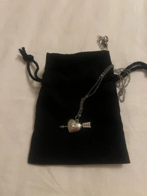 Silverfärgat halsband med hjärthänge - Snyggt halsband i silverfärg med ett hjärthänge som har en pil genom sig. Kedjan är tunn och smidig, perfekt för att ge en romantisk touch till din outfit.