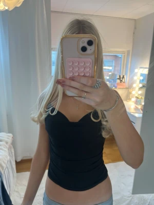 Svart croppad linnetopp - Snygg svart croppad linnetopp med smala axelband och v-ringad urringning. Perfekt att styla med jeans eller kjol för en trendig look. Tunn och mjuk i materialet, sitter tight och framhäver figuren. 