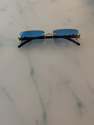 Blå rektangulära solglasögon med gulddetaljer - Snygga solglasögon med rektangulära blåtonade glas och guldfärgade detaljer, inklusive en leopardfigur på skalmarna. Glasögonen har minimalistisk båge och svarta skalmar för en modern look.