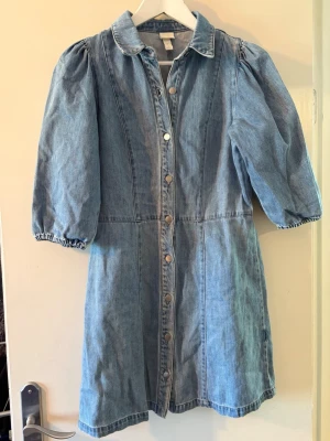 Jeansklänning med puffärm och knappar - Snygg jeansklänning i ljusblå denim med puffärmar och knappar hela vägen framtill. Klänningen har krage och trekvartsärm med resår vid ärmslut. Perfekt för dig som gillar en avslappnad men trendig stil.