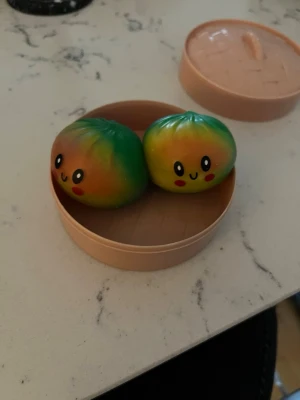Secret dumpling - Två squishies formade som dumplings med glada ansikten, i regnbågsfärger som grön, gul, orange, blå och rosa. De ligger i en beige plastkorg med lock, inspirerad av en ångkorg för dim sum. Perfekt som stressleksak eller dekoration. Jag kan även tejpa igen burken om du vill surprisea någon det är bara att säga till!!😊😊