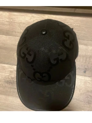 Svart keps från Gucci med GG-mönster -  Svart stilren keps från Gucci med det klassiska GG-mönstret som passar bra med varje outfit. Kepsen sitter otroligt bra på huvudet och går även att justera hur du vill ha den sittandes. I väldigt bra skick, knappt använd och inte använt den på över ett halvår