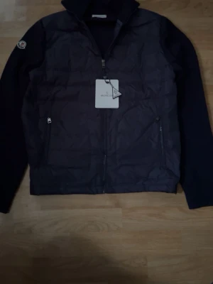 Mörkblå cardigan jacka från monclear  - Stilren mörk blå dunväst från Moncler med klassisk ståkrage och dragkedja framtill. Västen har två sidofickor med dragkedja och Moncler-logga på bröstet. Perfekt för lager-på-lager och håller dig varm med lätt dunfyllning.