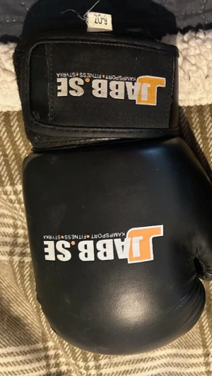 Svarta boxningshandskar JABB.SE barn - Ett par svarta boxningshandskar från JABB.SE med vit och orange logga på ovansidan. Handskarna har kardborreknäppning vid handleden och är tillverkade i syntetmaterial. Perfekta för kampsport eller träning. 6 Oz 