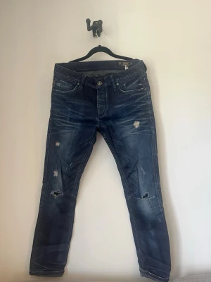 Blå slitna jeans med hål - Säljer ett par blå jeans från Jack and Jones originals med slitningar och hål på knäna. Jeansen har en slim fit passform och klassisk femficksdesign. Perfekta för en avslappnad och trendig look. Materialet är denim i bomull. Skick 7/10 storlek S/M
