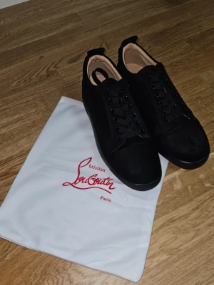 Svarta Christian Louboutin sneakers - Snygga svarta sneakers från Christian Louboutin med klassisk röd sula och diskret logga. Skorna har lågt skaft, svarta skosnören och ovandel i mocka och läder. Perfekta för dig som vill ha en stilren och exklusiv look. Storlek 43 och finns 44