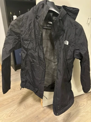 Svart vindjacka från The North Face - Snygg svart vindjacka från The North Face med huva och dragkedja. Jackan har meshfoder på insidan för extra ventilation och logga på bröstet. Perfekt för blåsiga dagar och enkel att packa ner i väskan.