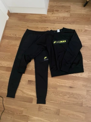 Nike Air Max svart loungewear-set - Svart loungewear-set från Nike Air Max med matchande sweatshirt och byxor. Båda plaggen har neongula Nike-loggor och Air Max-tryck. Mjukt material och avslappnad passform, perfekt för chill dagar hemma. 