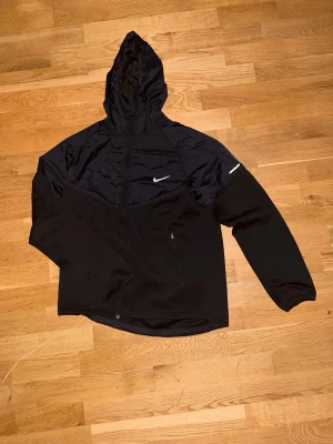 Svart Nike vindjacka med huva - Snygg svart vindjacka från Nike med huva och dragkedja. Jackan har reflekterande Nike-logga på bröstet och ryggen samt en mindre reflexdetalj på ärmen. Tillverkad i lätt och vindtätt material, perfekt för blåsiga dagar.