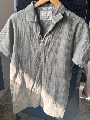 Grå ribbad polotopp med half zip - Snygg grå polotopp från Zara Knit med korta ärmar och ribbad struktur. Toppen har en halv dragkedja framtill och klassisk krage som ger en sportig vibe. Perfekt till jeans eller kjol för en trendig look.