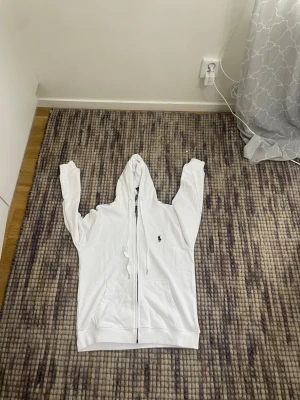 Vit hoodie från Ralph Lauren - Säljer en stilren vit hoodie från Ralph Lauren med dragkedja framtill och klassisk huva. Tröjan har två fickor och den ikoniska mörkblå polospelaren broderad på bröstet. Perfekt för dig som gillar enkel och tidlös design.