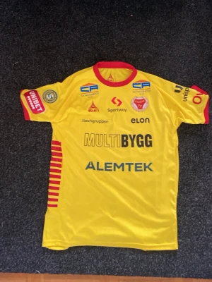 Gul matchtröja Kalmar FF nr 28 - Säljer en gul matchtröja från Kalmar FF med röda detaljer och flera sponsorer tryckta på både fram- och baksida. Tröjan har nummer 28 och namnet Sabriye på ryggen. Aldrig använd. Pris går att diskutera. 