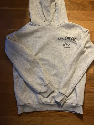 Ljusgrå hoodie med tryck Ma Chérie - Ljusgrå hoodie med svart tryck 'MA CHÉRIE' och en krona både fram och bak. Mjuk och skön med ribbade muddar och klassisk huva. Perfekt för en avslappnad och trendig stil.