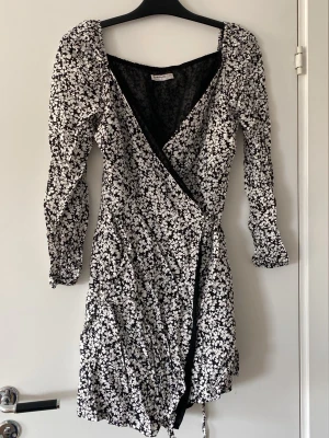 Svartvit omlottklänning från ASOS - Snygg svartvit omlottklänning från ASOS med blommigt mönster. Klänningen har v-ringning, långa ärmar och knyt i midjan.