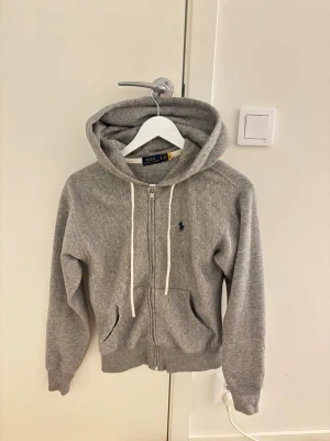 Grå zip hoodie från Polo Ralph Lauren - Klassisk grå hoodie med dragkedja från Polo Ralph Lauren. Tröjan har huva med vita snören, två fickor framtill och den ikoniska blå loggan broderad på bröstet. Väldigt mjukt och skönt material!