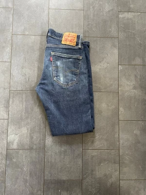 Levi's 514 raka blå jeans - Säljer ett par klassiska Levi's 514 jeans i blå denim med raka ben och normal midja. Jeansen har en bakficka med den ikoniska röda Levi's-lappen och slitna detaljer för en cool vintagekänsla. Perfekta till vardagslooken!