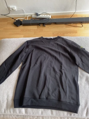 Stone island tröja - Svart långärmad sweatshirt från Stone Island med klassisk rund hals och ribbade muddar. Tröjan har den ikoniska loggan på ärmen och är tillverkad i mjukt bomullsmaterial. Perfekt för en stilren och avslappnad look.