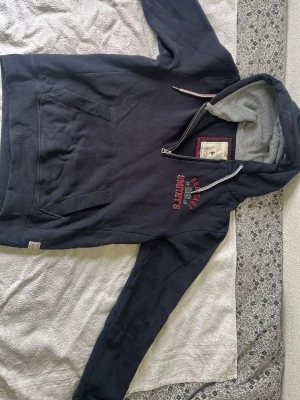 Mörkblå hoodie från Smithy's England - Mörkblå hoodie från Smithy's England med broderad text och Union Jack-detalj på bröstet. Halvlång dragkedja, stor känguruficka och fodrad huva med dragsko. Perfekt för en avslappnad och stilren look.