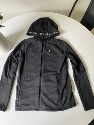 Svart hoodie från Peak Performance -  Pris går att diskutera Snygg svart hoodie från Peak Performance med dragkedja framtill och två fickor med dragkedja. Huva med logotyptext på insidan och liten logga på bröstet. Tillverkad i mjukt och stretchigt material, perfekt för en sportig och avslappnad stil.