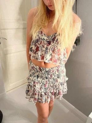 Blommig topp linne zara trend snygg - Söt croppad linnetopp med blommigt mönster i beige, lila och vinröda toner. Toppen har tunna axelband, v-ringning och volangig nederkant. Perfekt för varma dagar och somriga outfits.