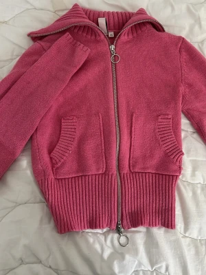 Rosa ziptröja från Nelly - Säljer en rosa stickad ziptröja från Nelly med två framfickor och bred ribbstickad mudd vid ärmslut och nederkant. Tröjan har en hög krage och dragkedja med ringdetalj. 