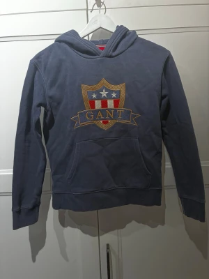 Blå hoodie från GANT med emblem - Mörkblå hoodie från GANT med stort broderat emblem på bröstet i rött, vitt och guld. Klassisk känguruficka framtill och huva. Mjuk bomullskänsla och sportig look, perfekt för dig som gillar preppy stil. Nypris (1100)