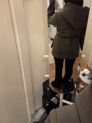Kappa  - Snygg mörkgrå kappa med huva och knytskärp i midjan. Kappan har en avslappnad passform och är tillverkad i ett mjukt tyg som ger en stilren look. Perfekt för kyligare dagar och enkel att matcha med olika outfits.