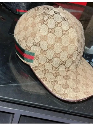 Beige keps från Gucci med GG-mönster - Snygg beige keps från Gucci med klassiskt GG-monogram över hela kepsen. Har gröna och röda ränder på sidan som ger en ikonisk touch. Tillverkad i canvasmaterial och har böjd skärm. Perfekt för dig som vill sticka ut med en lyxig accessoar.
