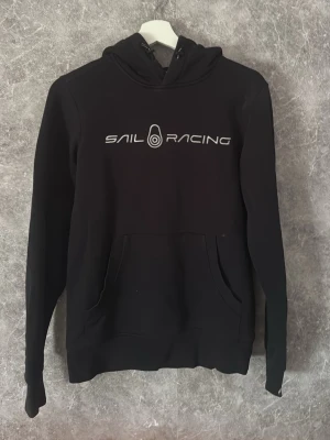  hoodie Sail Racing - Svart hoodie med Sail Racing-logga i vitt på bröstet. Klassisk modell med huva och känguruficka framtill. Perfekt för en avslappnad och sportig stil. Mjuk bomullskänsla och ribbade muddar vid ärmslut och nederkant.