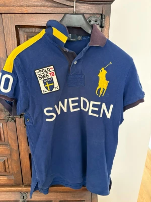 Marinblå Polo Ralph Lauren skjorta - Marinblå pikéskjorta från Polo Ralph Lauren med stora gula detaljer, broderad logga och texten SWEDEN framtill. Skjortan har korta ärmar, klassisk krage och knappar vid halsen. Materialet är bomull och polyester, perfekt för en sportig look inför fotbolls-VM. 