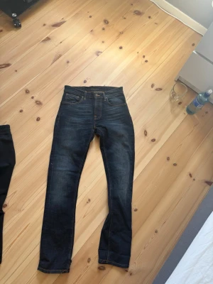 Mörkblåa jeans från Nudie - Snygga mörkblåa  jeans från Nudie med klassiska fem fickor och orangea kontrastsömmar. Jeansen har normal midja och raka ben. Tillverkade i denim med stretch för extra komfort. Perfekta för en trendig och avslappnad look. Jeansen är i nytt skick med noll defekter.