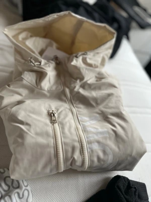 Beige vindjacka från monclair - Snygg beige vindjacka från Tommy Hilfiger med hel dragkedja, huva och diskret logga på bröstet. Jackan har en klassisk passform och är tillverkad i lätt, vindtätt material. Detalj med Hilfigers signaturrand på ärmen ger en stilren touch.