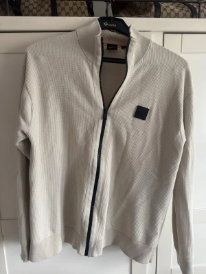 Beige ziptröja från BOSS - Stilren beige ziptröja från BOSS i relaxed fit. Tröjan har en diskret struktur, hel dragkedja framtill och en svart logopatch på bröstet. Perfekt för en avslappnad men snygg look.