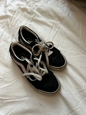 Svarta Vans Old Skool sneakers - Klassiska Vans Old Skool sneakers i svart canvas och mocka med vit sula och vit sidostripes. Skorna har lågt skaft, vita skosnören och den ikoniska gummisulan med rutmönster. Perfekta för en streetwear-look.