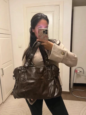 Mörkbrun läderhandväska med nitar - Snygg mörkbrun handväska i läder med dubbla handtag och dekorativa nitar. Väskan har en mjuk, något slouchy form och silverfärgade metalldetaljer. Väskan är i mycket bra skick!