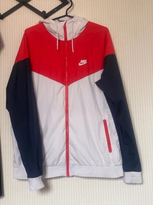 Nike vindjacka röd, vit och marinblå - Nike vindjacka med huva i rött, vitt och marinblått. Jackan har dragkedja framtill, två fickor med dragkedja och vit Nike-logga på bröstet. Lätt material, perfekt för blåsiga dagar. Sportig design med färgblock och justerbar huva.