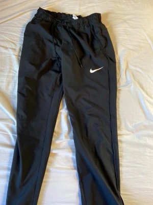 Svarta Nike mjukisbyxor med dragsko - Säljer ett par svarta mjukisbyxor från Nike med klassisk vit Swoosh-logga på benet. Byxorna har resår i midjan, dragsko och sidofickor. Perfekta för chill eller träning, tillverkade i mjukt och bekvämt material.