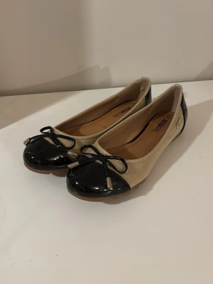 Ballerinaskor, svart och beige - Helt nya ballerinaskor med en glansig yta med rosetter. Säljer för att de var för små för mig, jag skulle säga att de funkar om man har storlek 38, den angivna storleken är 39. Pris kan diskuteras. 
