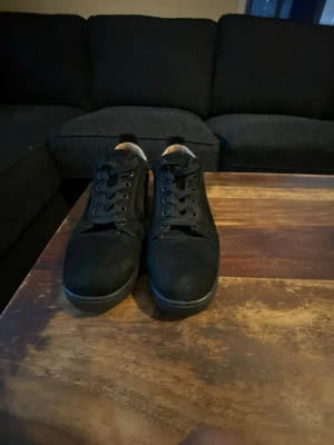 Svarta sneakers med röd sula - Svarta sneakers med klassisk låg profil och snörning. Skorna har en snygg röd yttersula som ger en cool kontrast. Ovandelen verkar vara i mocka eller liknande material. Perfekta för dig som gillar stilrena och diskreta sneakers med en twist.