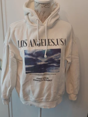 Vit hoodie med tryck Los Angeles, USA - Vit hoodie med stor huva och dragsko. På framsidan finns ett tryck med texten 'LOS ANGELES, USA' och en bild av en solnedgång samt texten 'FIND YOUR PERFECT SUNSET'. Mjuk insida och ribbade muddar vid ärmslut och nederkant.