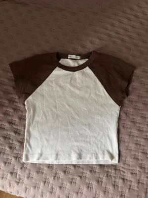 Brun och vit ribbad t-shirt - Snygg ribbad t-shirt från Bik Bok med bruna ärmar och krage samt vit kropp. Klassisk basebollstil med korta ärmar och rund hals. Perfekt till jeans eller kjol för en avslappnad look.